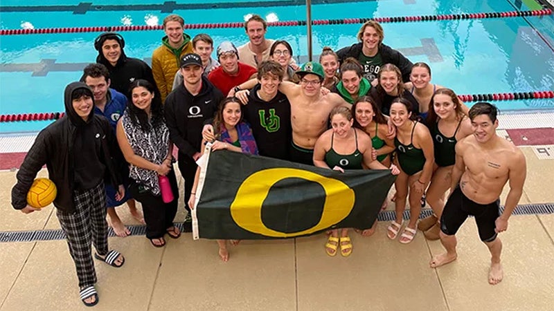 Male and female members of the Club Sports swimming team stand by the pool and hold a banner with the "O"