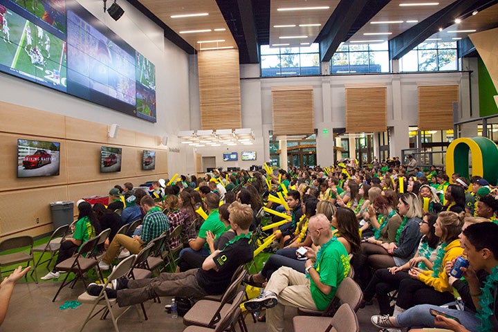 Lounge full of people sitting and watching a Ducks footbal game on the big screens.