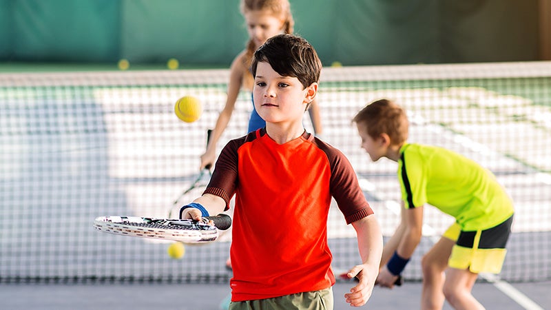 A young boy practices hitting the ball with his tennis racquet while other kids practice behing him.
