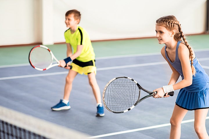 Two kids stand in the ready position with their tennis racquets waiting for the tennis ball to come their way.
